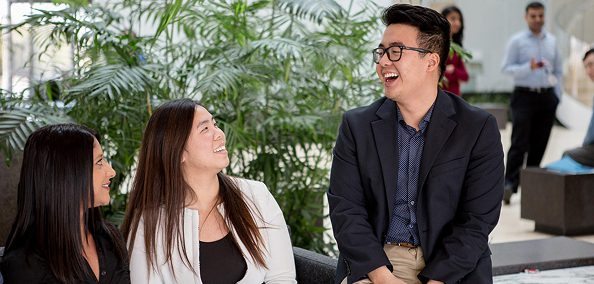 Three coworkers smiling and chatting indoors, seated near plants in a bright office space.