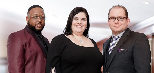 Three diverse professionals posing together, smiling at the camera in formal business attire.